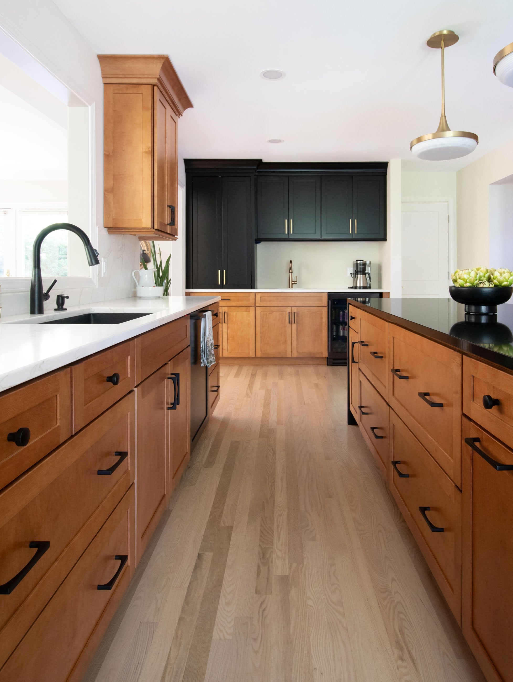 Warm kitchen remodel featuring shaker cabinetry with warm wood stain and black accents, with black quartz waterfall countertop on island - galley view with beverage center at rear