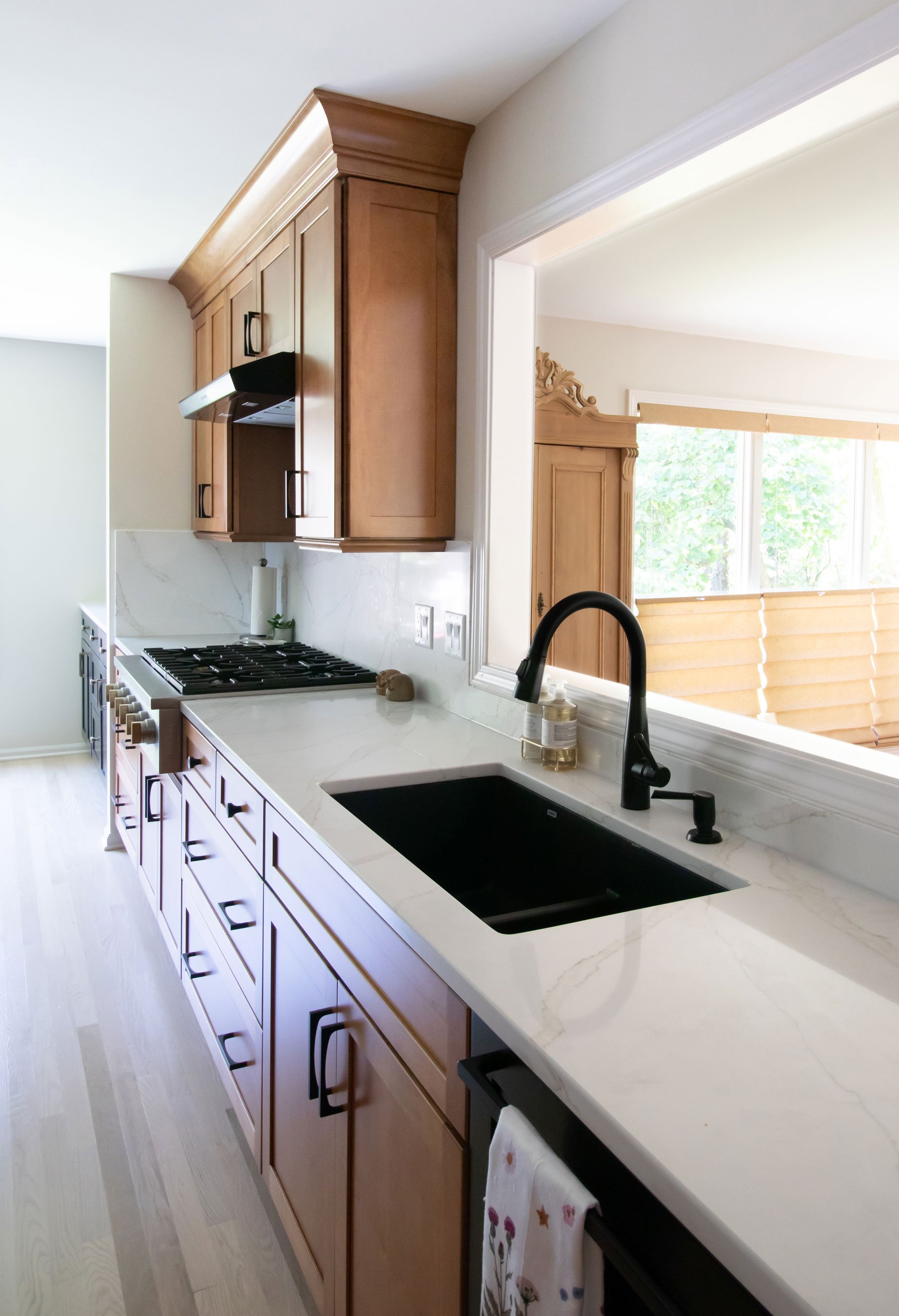 Warm kitchen remodel featuring shaker cabinetry with warm wood stain and black accents, with black quartz waterfall countertop on island - view showing sink and pass-through to living room